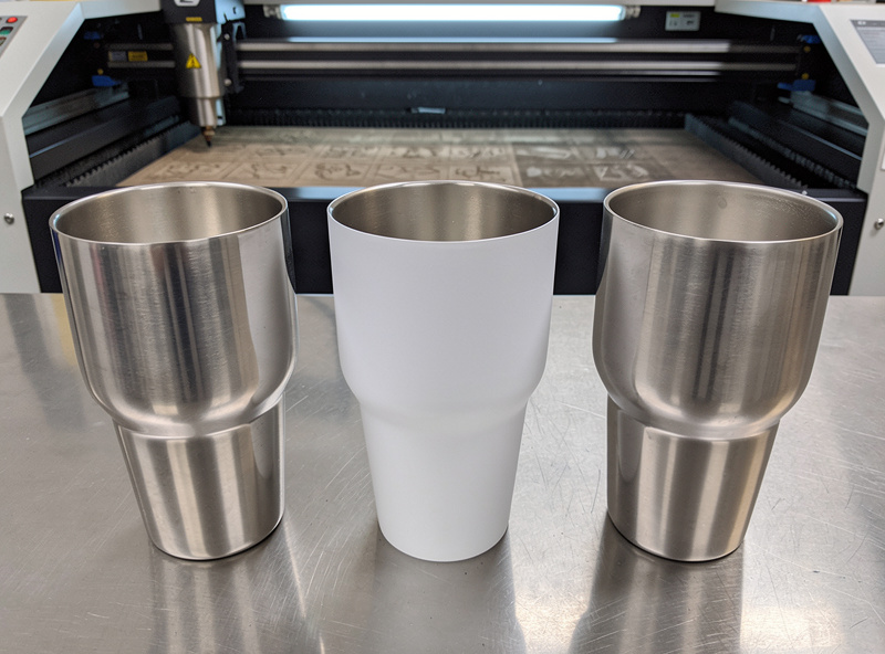 Top down view of stainless steel tumblers prepared for laser engraving with Cermark spray coating applied to one tumbler and a CO2 laser nearby