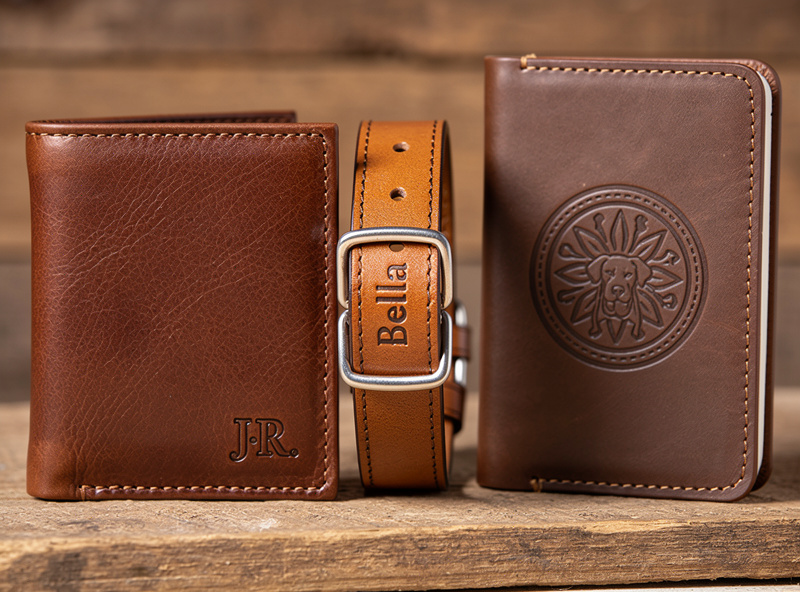Three finished laser engraved leather items on a wooden display: a wallet with monogrammed initials, a dog collar with engraved name, and a journal cover with logo all in vegetable tanned leather