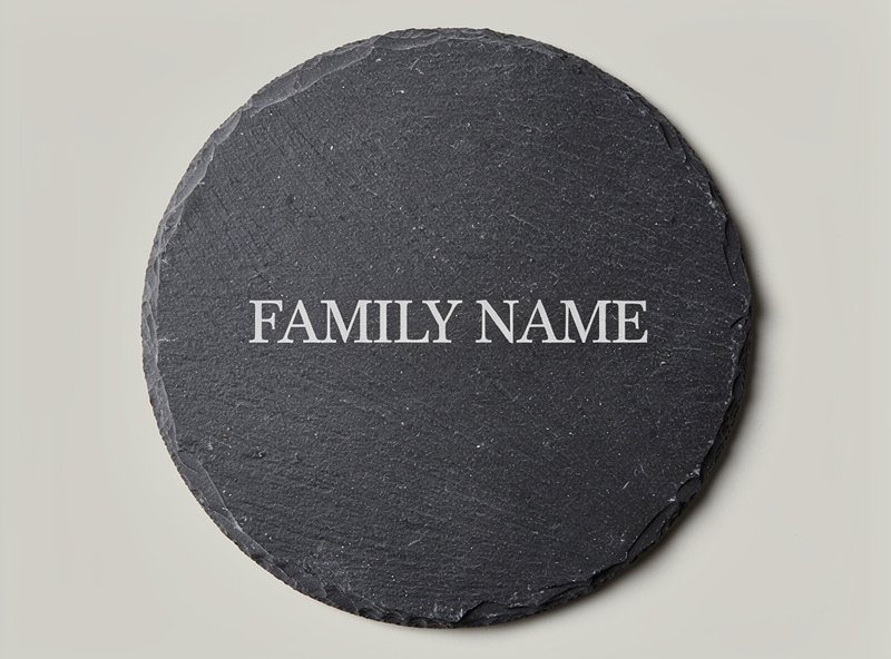 Round slate coaster with white laser engraved family name and decorative border design centered on the dark gray slate surface
