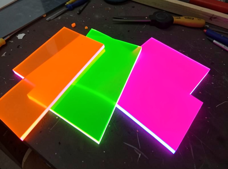 Fluorescent acrylic offcuts with glowing coloured edges arranged on a dark workbench, the cut edges appearing to emit light