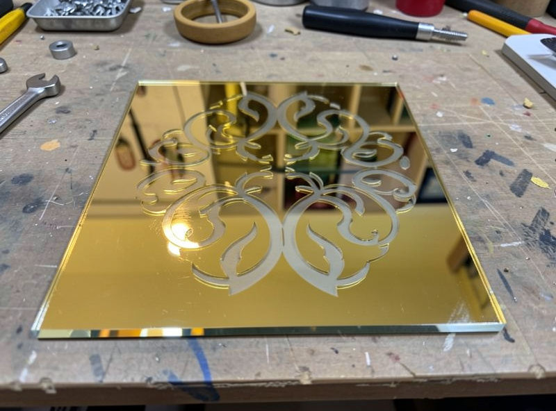 A piece of mirrored gold acrylic with a design engraved through the mirror coating to reveal the clear base underneath, on a workbench