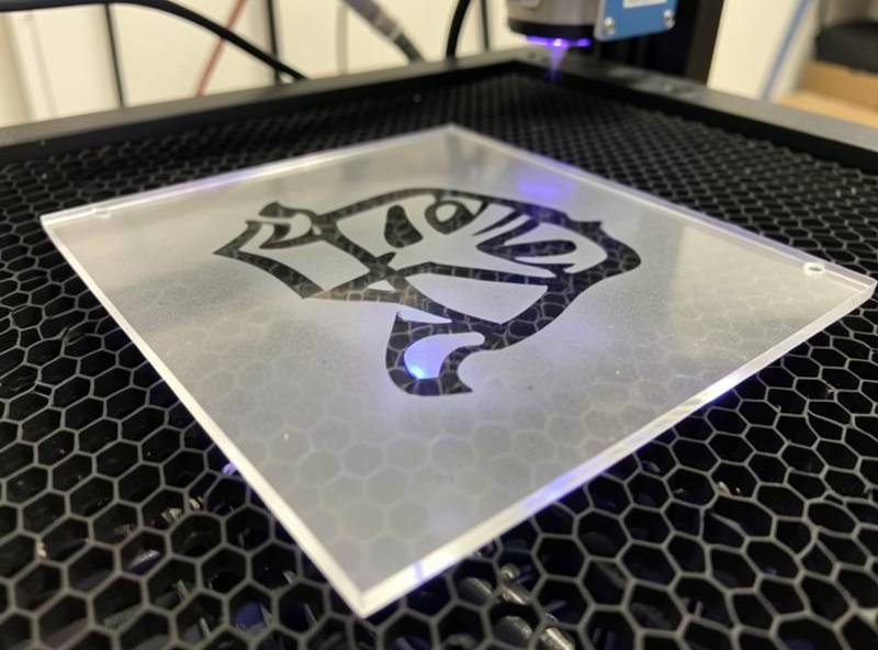 Close-up of a clear acrylic panel on a laser bed mid-engrave with a frosted fill pattern forming the design that will later be edge-lit
