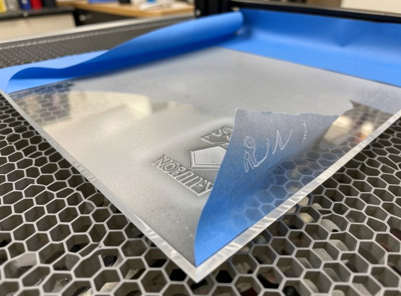 A sheet of cast acrylic on a laser honeycomb bed with the paper protective film partially peeled back to show a freshly engraved area