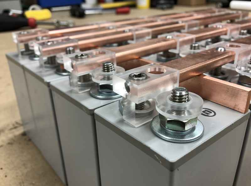 Laser-cut clear acrylic bus bar insulators installed between copper bus bars and LiFePO4 battery terminals, clean DIY battery bank build on workbench