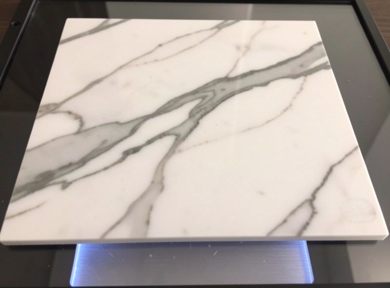A white marble tile on a laser bed with a softly darkened etched design, natural grey veining visible across the surface