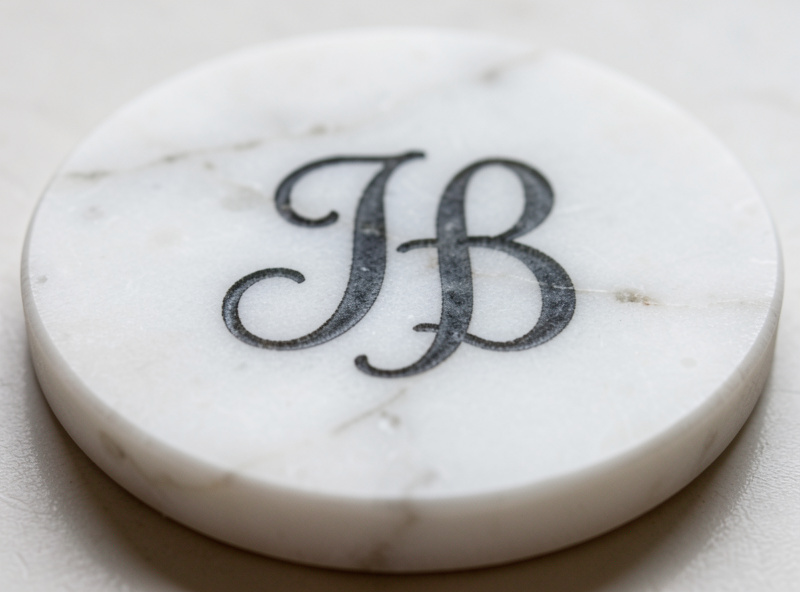 Close-up of a softly etched darkened monogram on a polished white marble coaster with natural veining