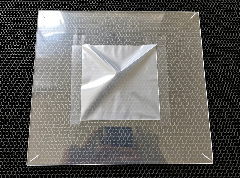 A clear acrylic sheet on a laser bed with masking film peeled away only over the central engraving area while the surrounding film stays in place