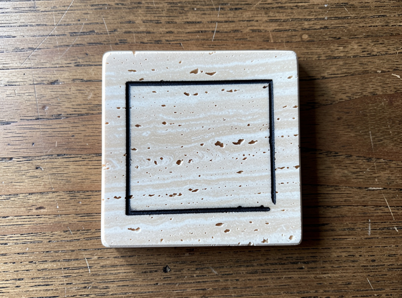 A finished sealed travertine coaster with a dark paint-filled engraved design on a wooden table