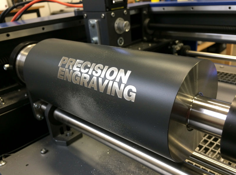 A powder-coated stainless tumbler mounted on a laser rotary attachment mid-engrave, revealing bright metal lettering